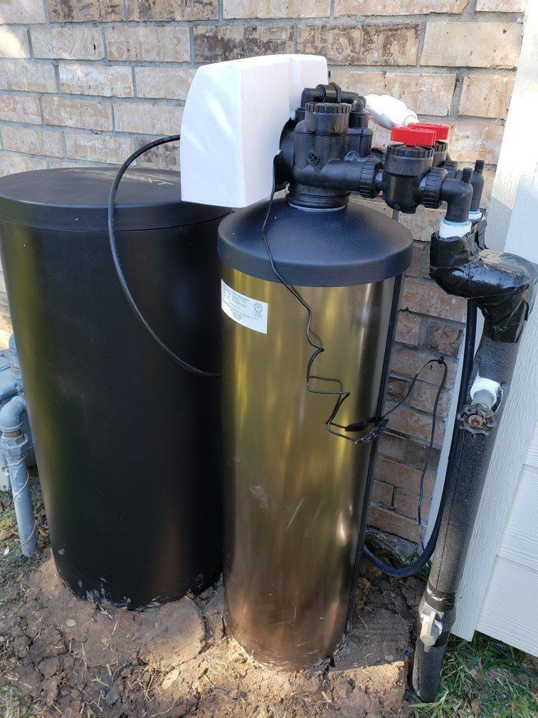 Outdoor water filtration equipment with two large black and metallic tanks beside a brick wall.