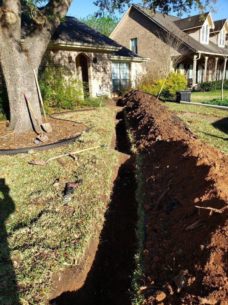 Long trench dug through a front yard beside a house, with exposed soil and a large tree nearby.