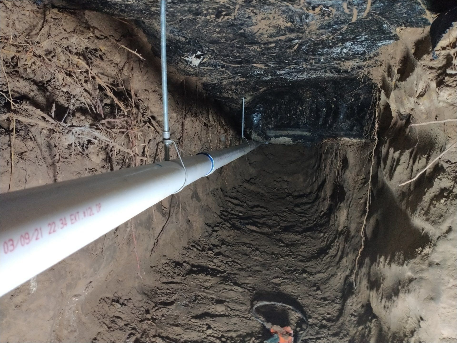 Narrow underground trench with a white pipe running along the left wall and rough dirt walls and floor.