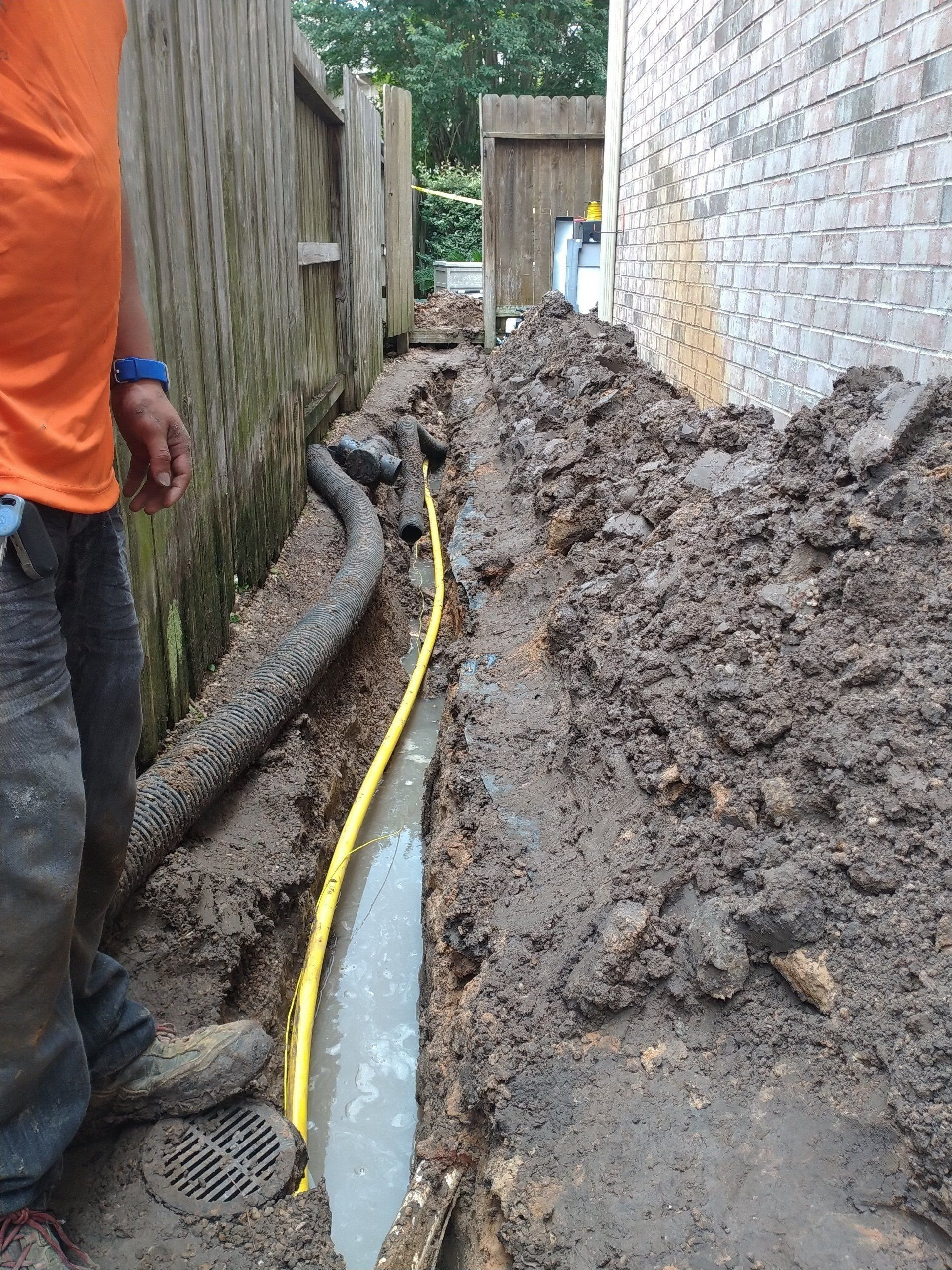 Narrow muddy trench with a yellow cable and workers beside a stone wall and fence