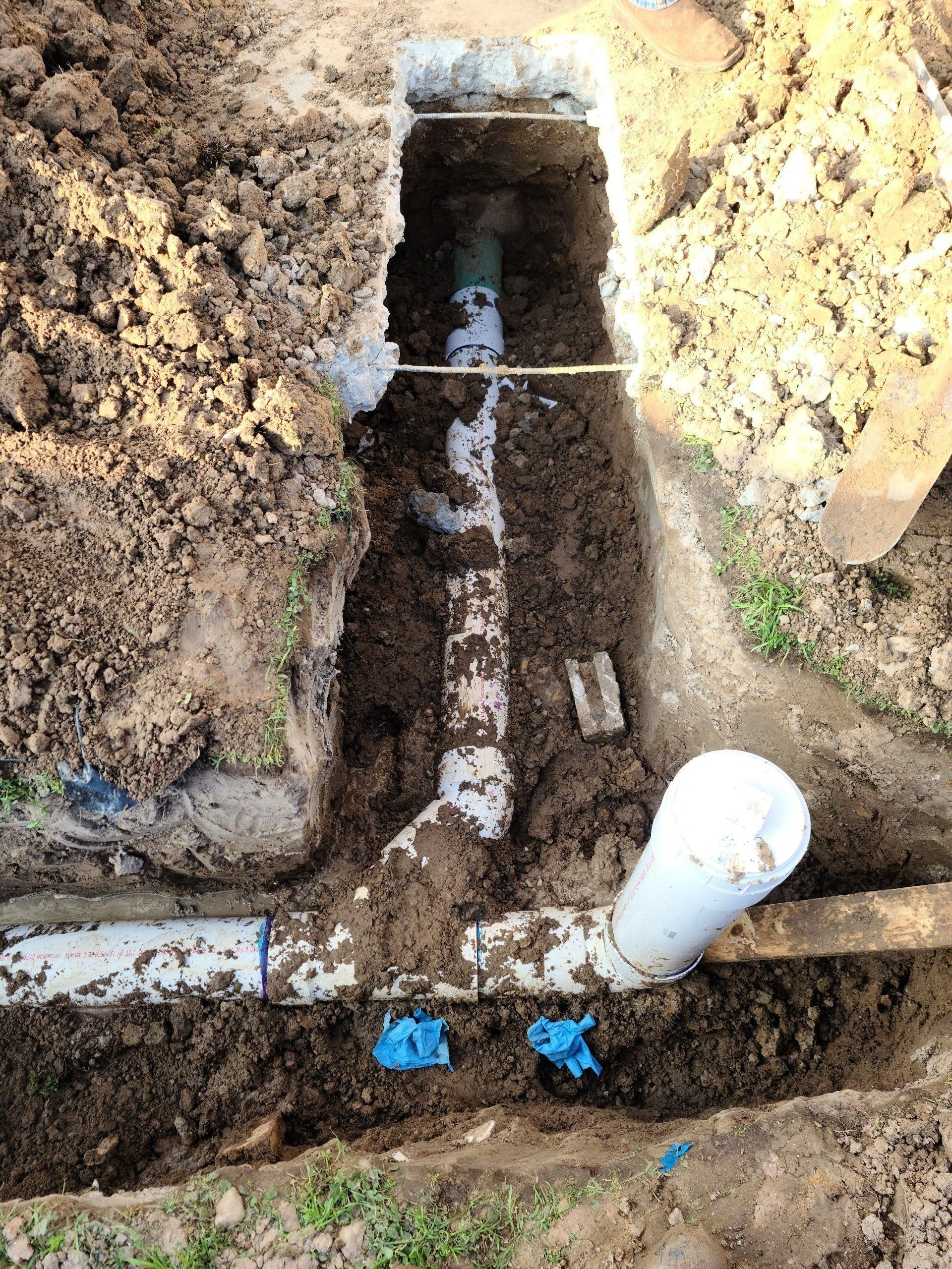 Utility trench with exposed pipes, dirt walls, and a white PVC pipe connection at ground level