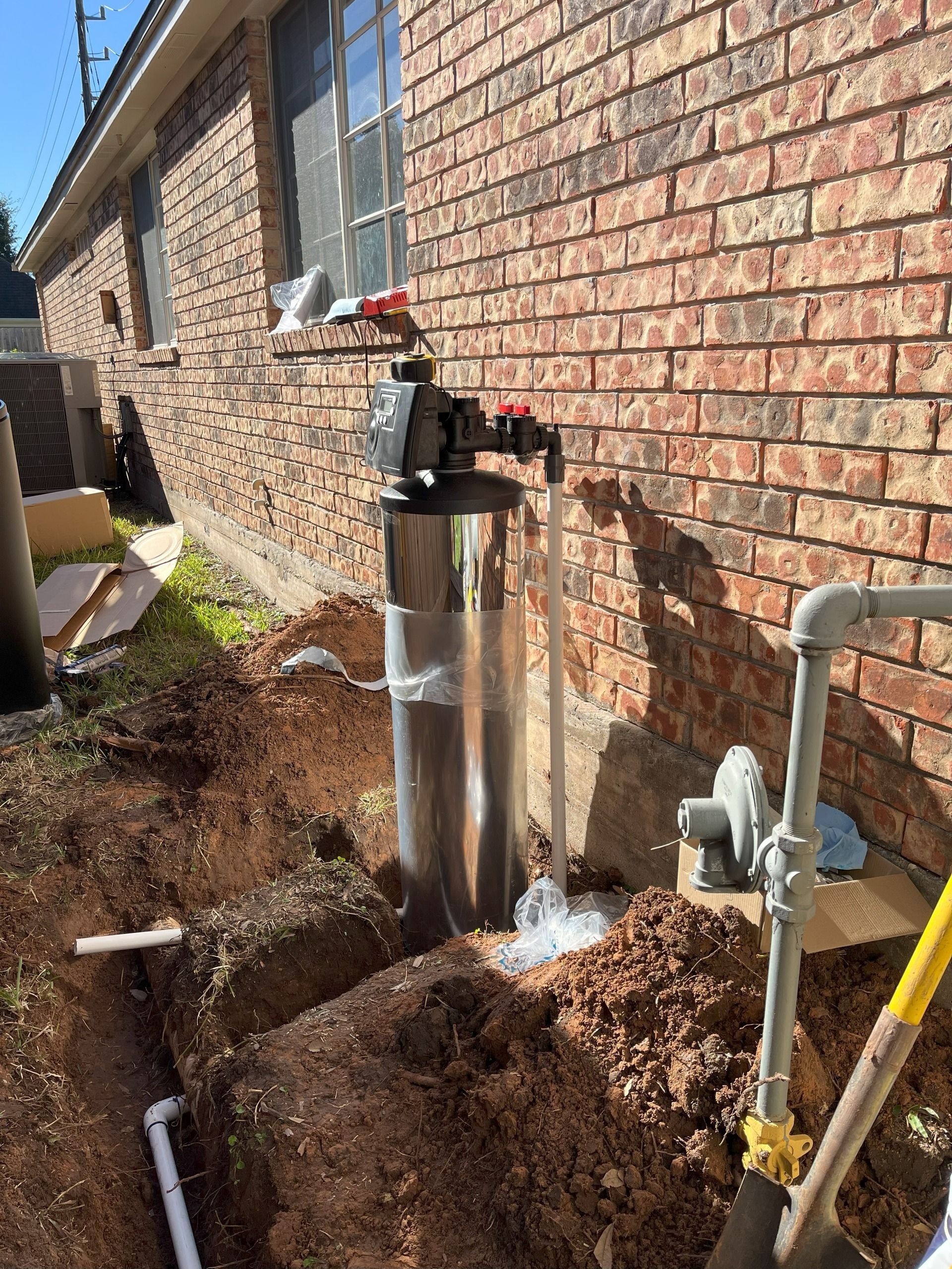 Shiny metal water heater and pipes beside a brick house, with a dug-up trench and soil piled nearby.