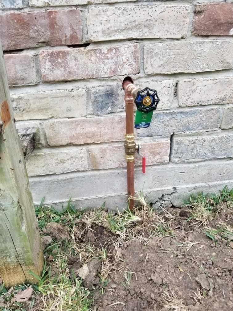 A brass outdoor water valve with a meter against a brick wall beside bare soil and grass.