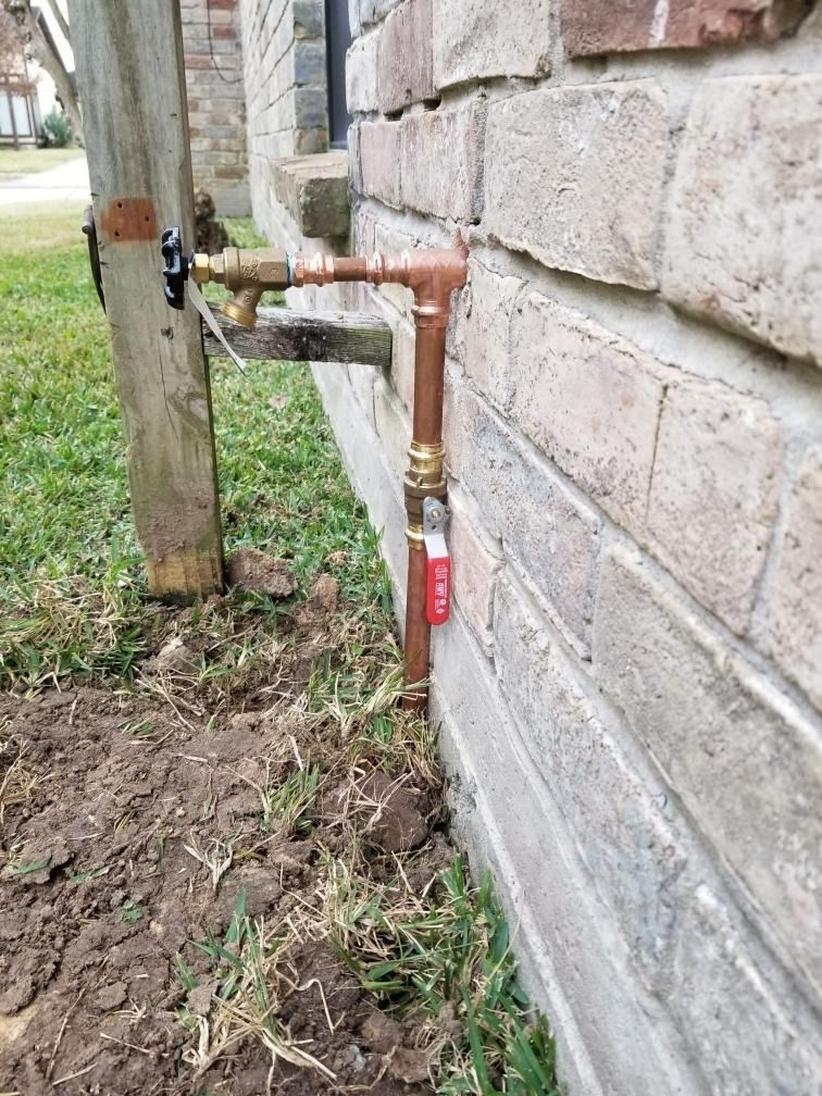 Copper pipe and red shutoff valve on a stone house wall near a wooden post outdoors.