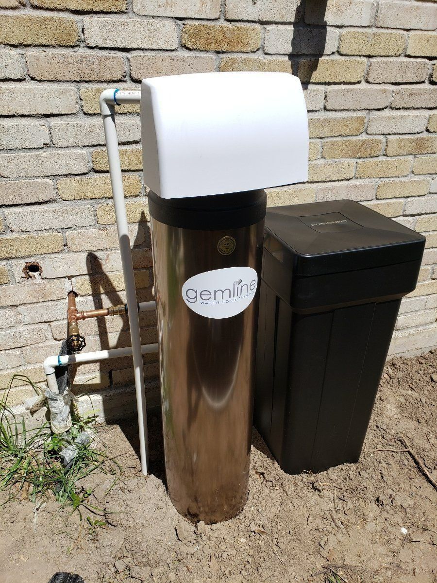 Outdoor white-and-black utility unit beside a brick wall and black storage bin on dirt ground