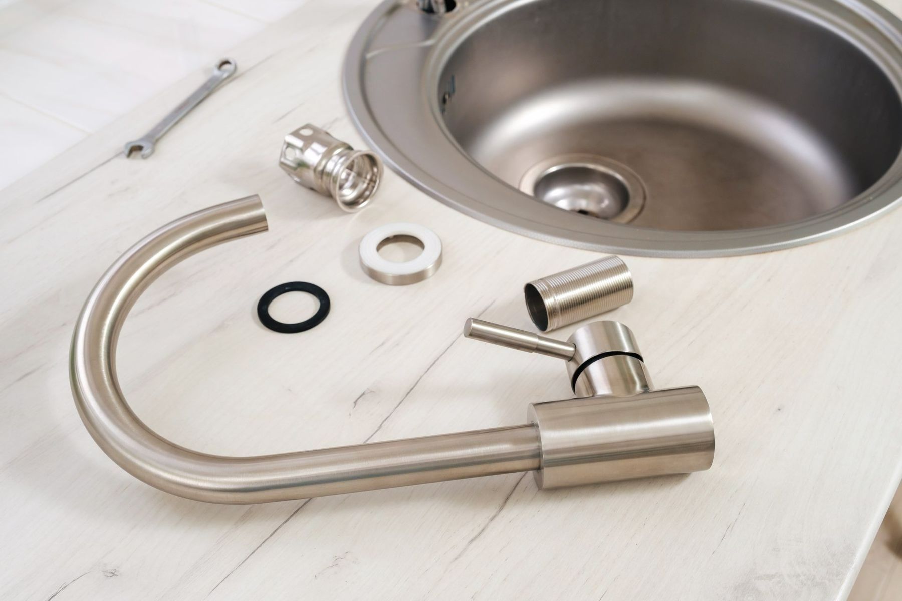 Kitchen sink faucet components laid out on a white countertop beside a stainless-steel sink.