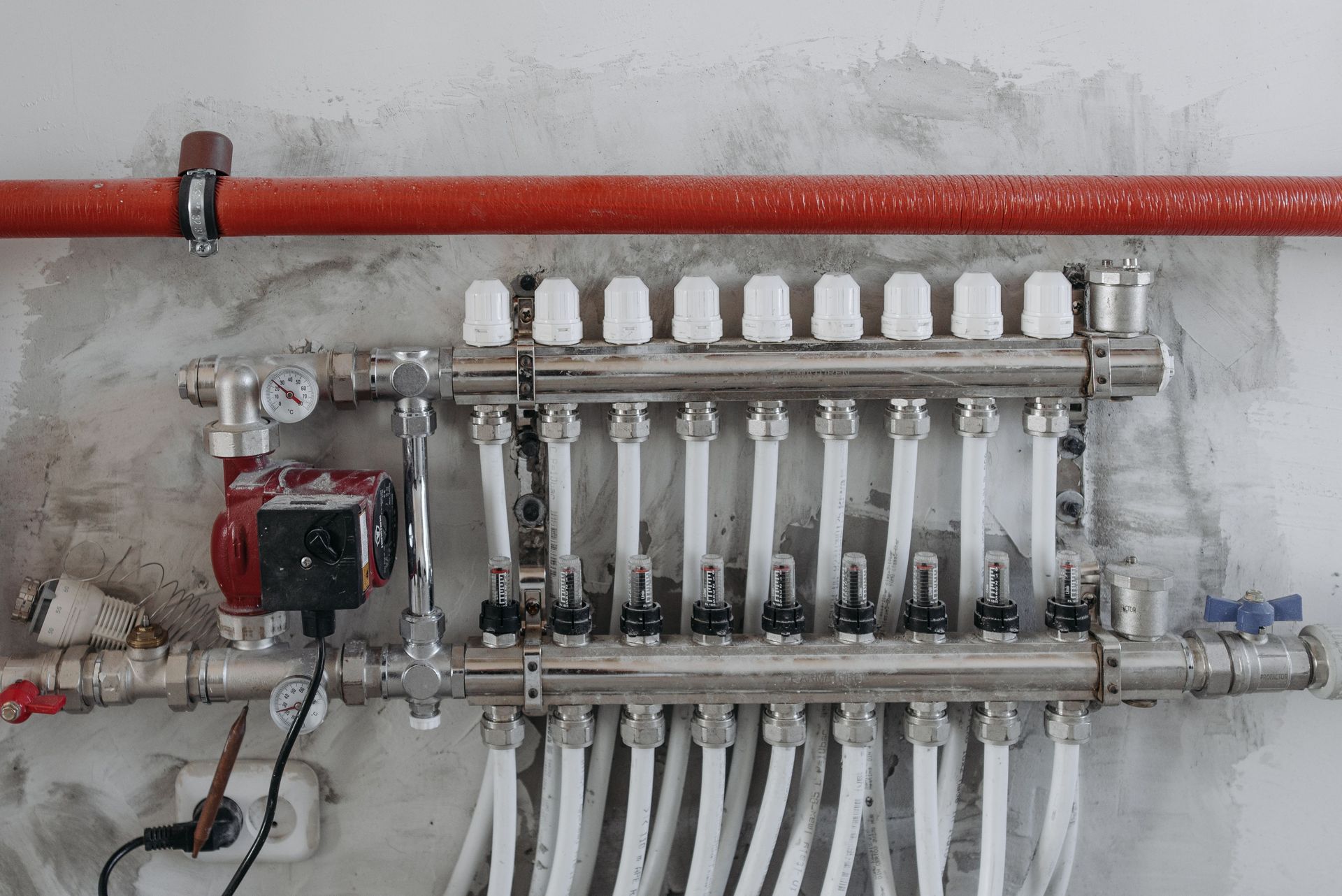 Plumbing manifold with multiple white valves and pipes mounted on a concrete wall, with a red pipe above.