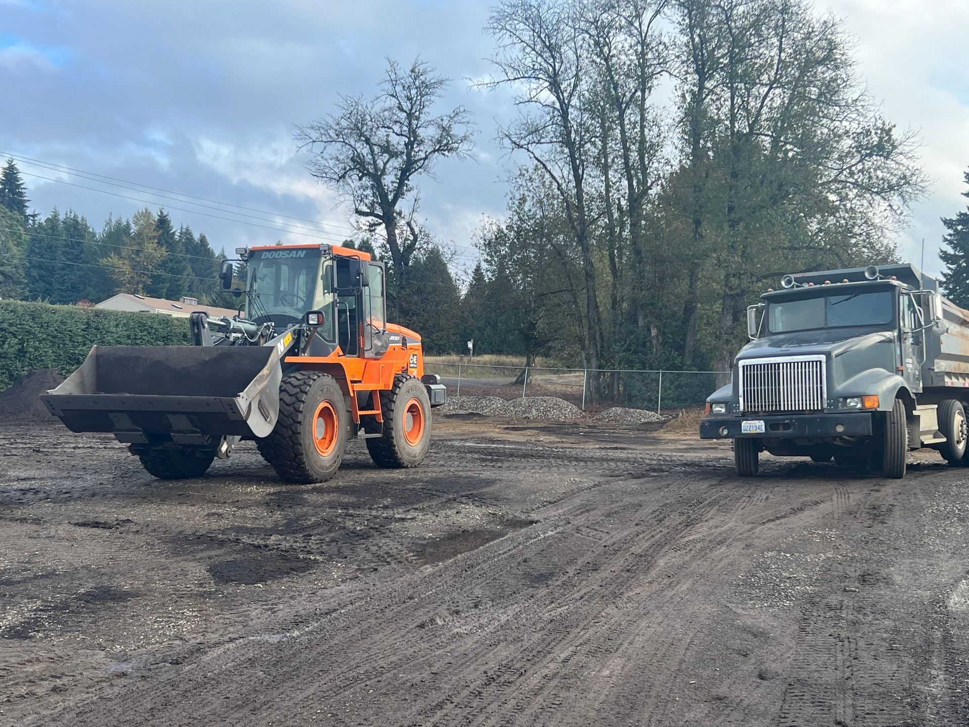 Construction site with excavator loading dirt into a dump truck; houses under construction in the background.