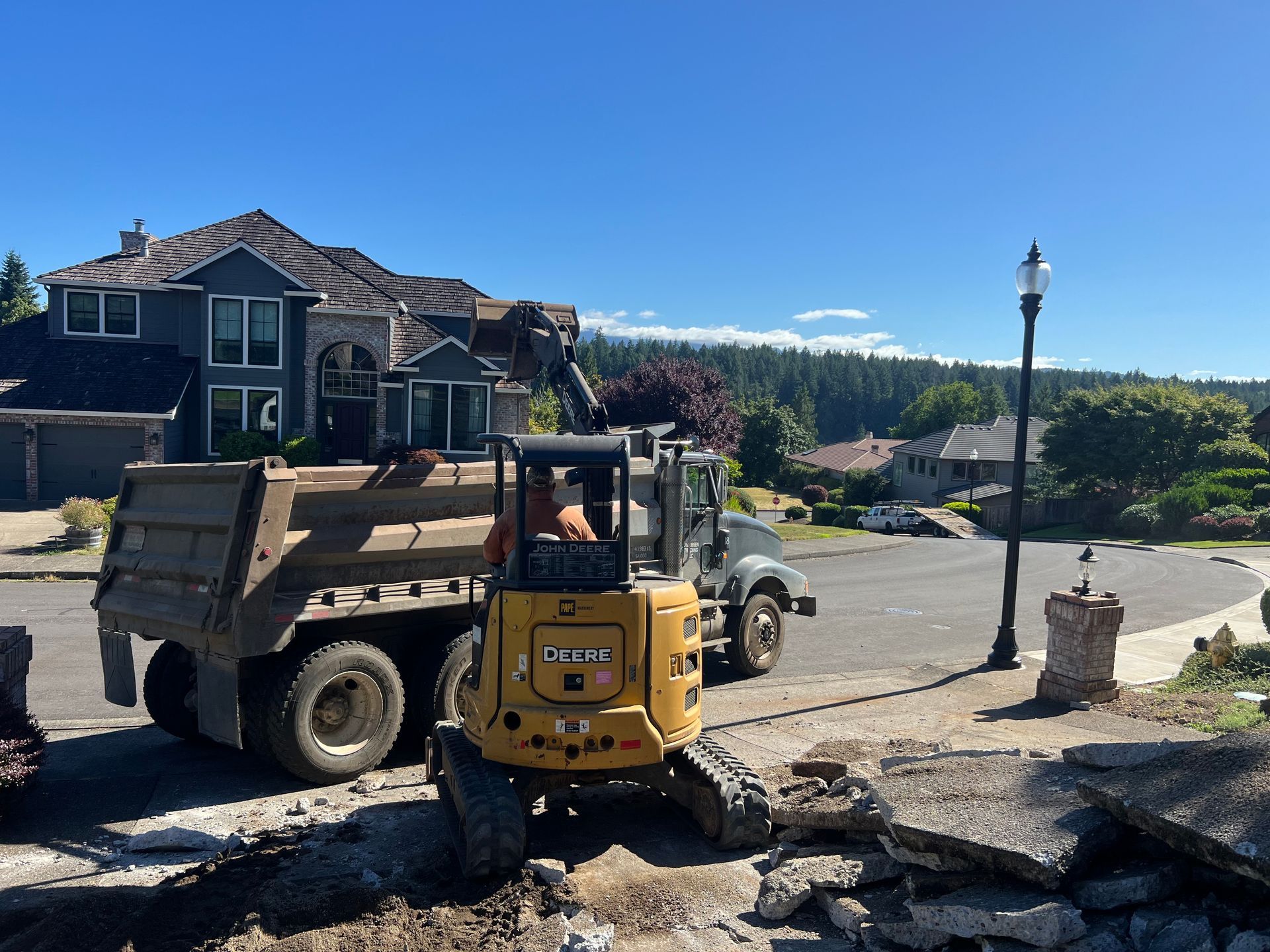 Red excavator digging on a city street. It's parked on a paved area next to a pile of dirt. White buildings are in the background.