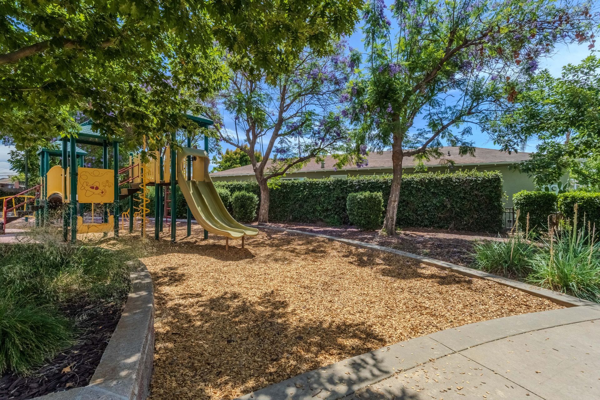 Shaded playground with slides and climbing structures beside a paved path and hedges