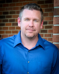 A man in a blue shirt is standing in front of a brick wall.