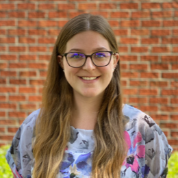 Paralegals Richmond VA: A woman wearing glasses and a floral shirt is smiling in front of a brick wall.