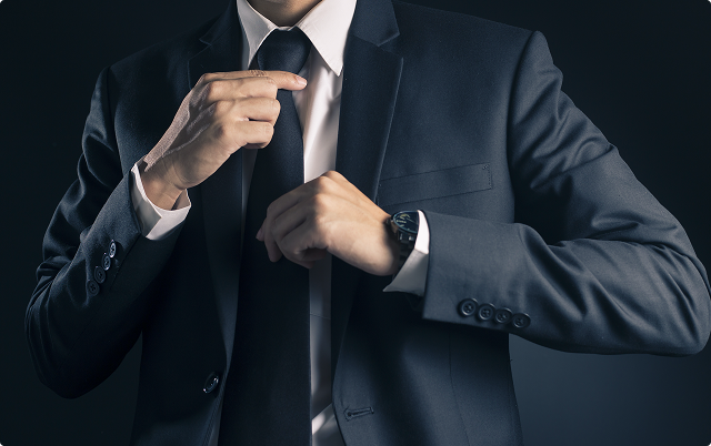 Man in a dark suit and tie adjusting his tie against a dark background.