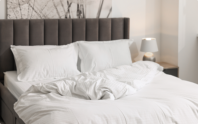 A bed with white bedding and pillows, a gray headboard, and a lamp on the nightstand.
