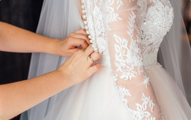 Hands buttoning up the back of a white wedding dress with lace sleeves.