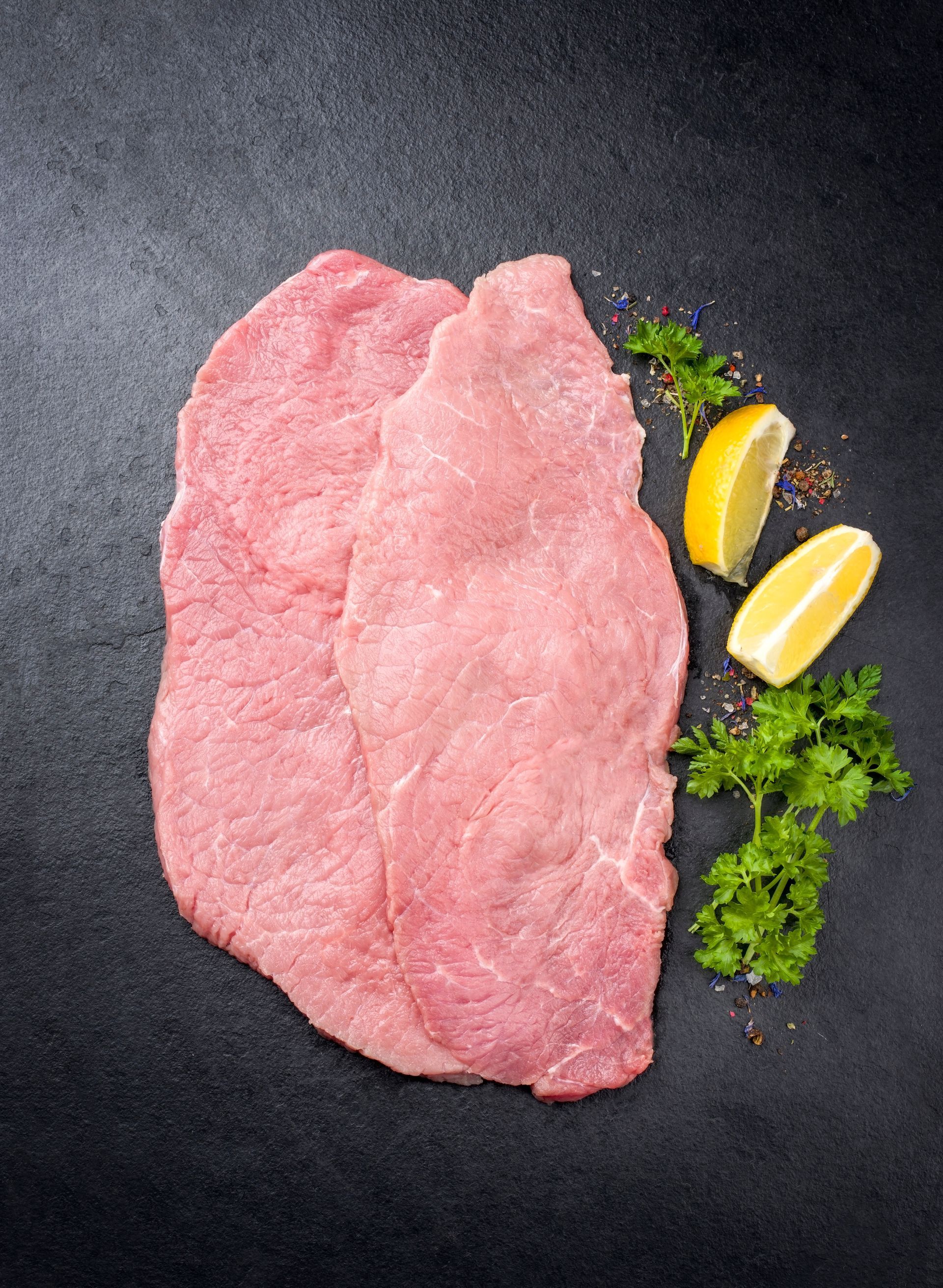 Dünne rohe Fleischschnitzel mit Zitrone und Petersilie auf dunklem Untergrund.