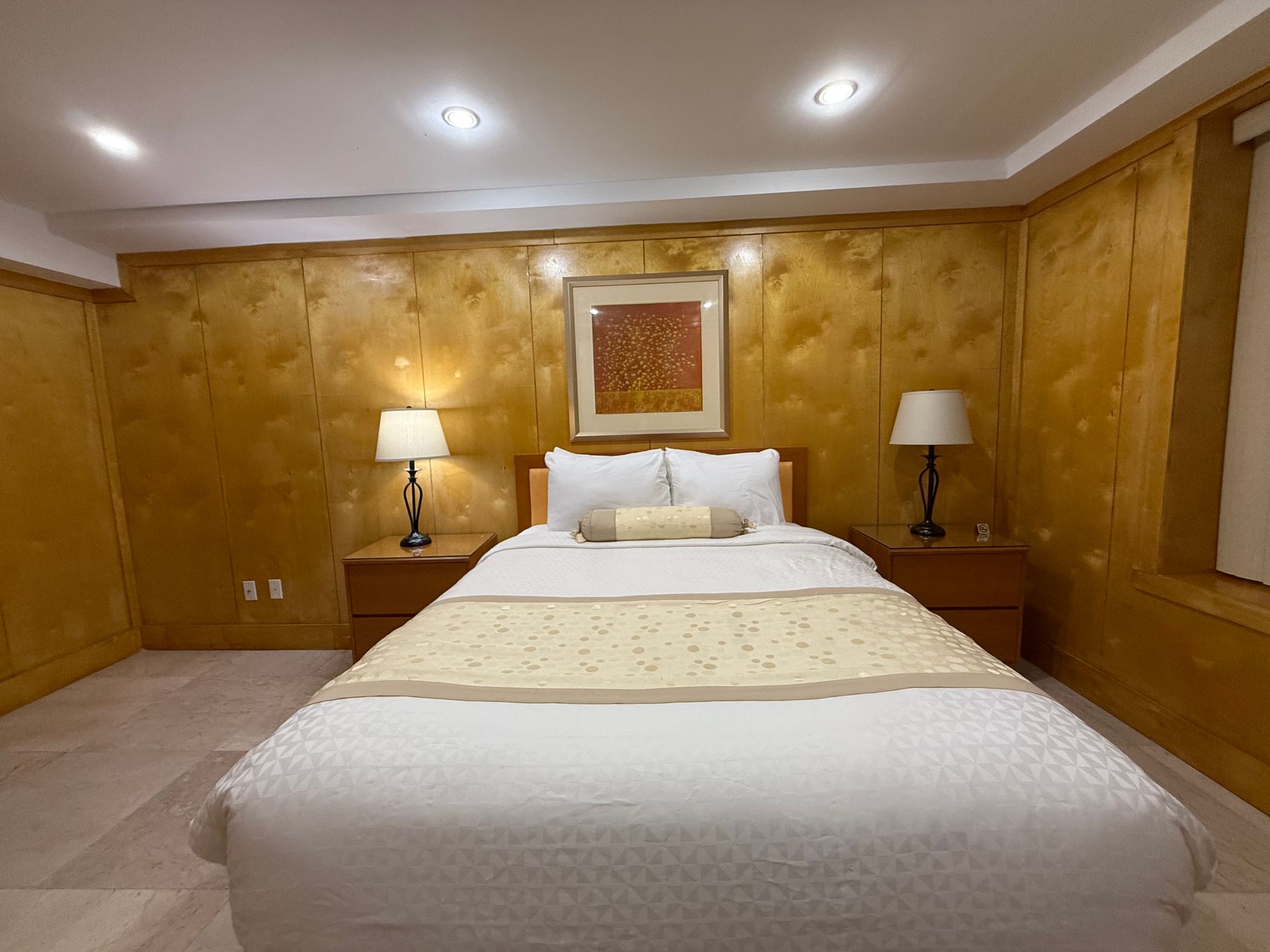 Bedroom with a bed, nightstands, lamps, and a framed artwork on a textured, wood-paneled wall.