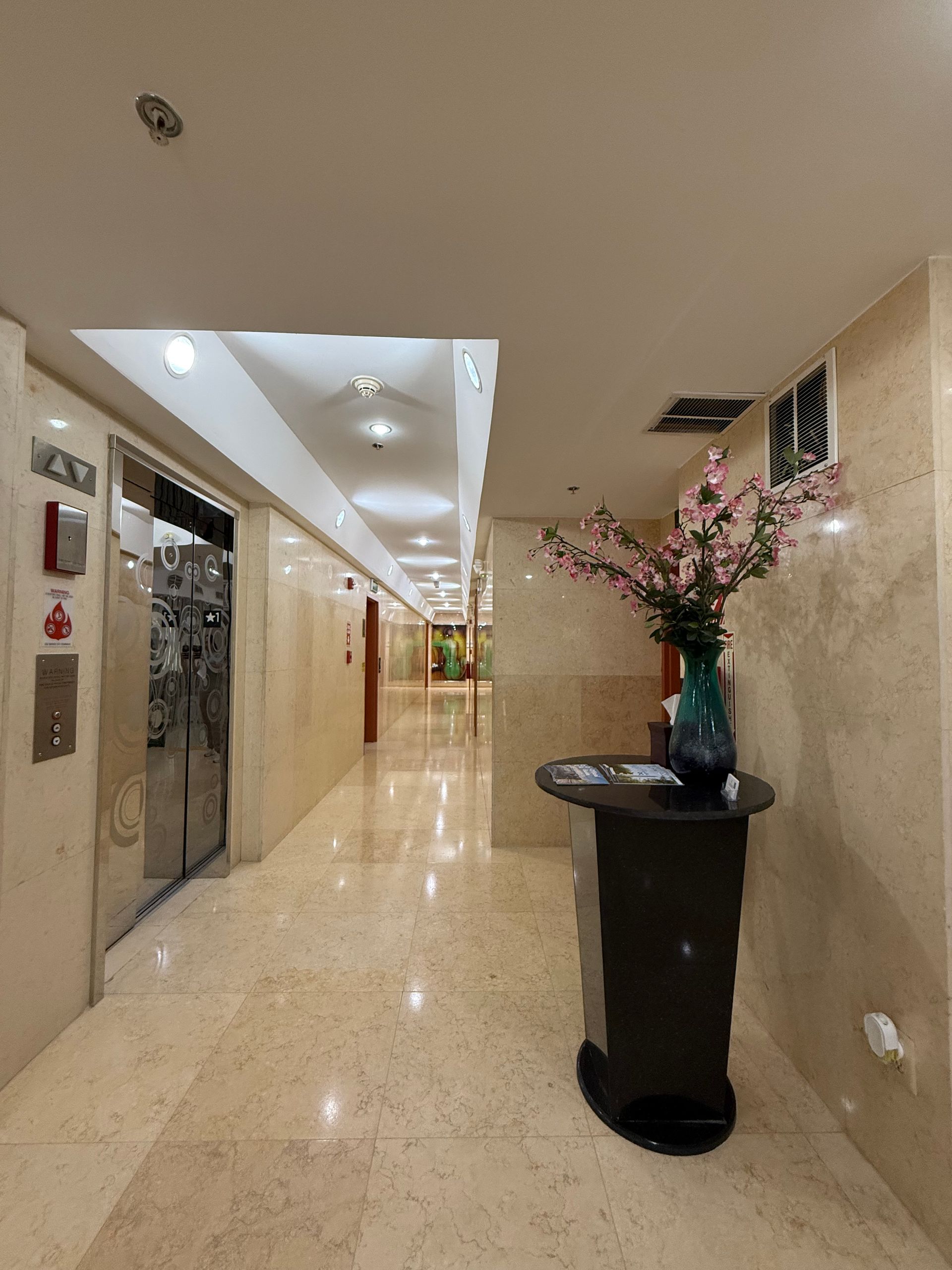 Long hallway with marble floors, elevator, and a vase of pink flowers.
