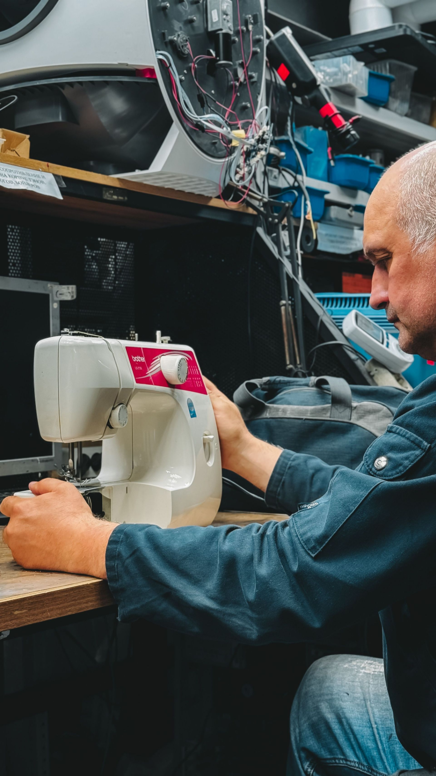 Man repairing a white sewing machine at a workbench in a cluttered workshop