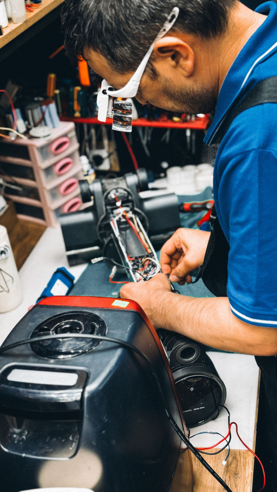 Person repairing a black car side mirror with tools in a cluttered workshop.