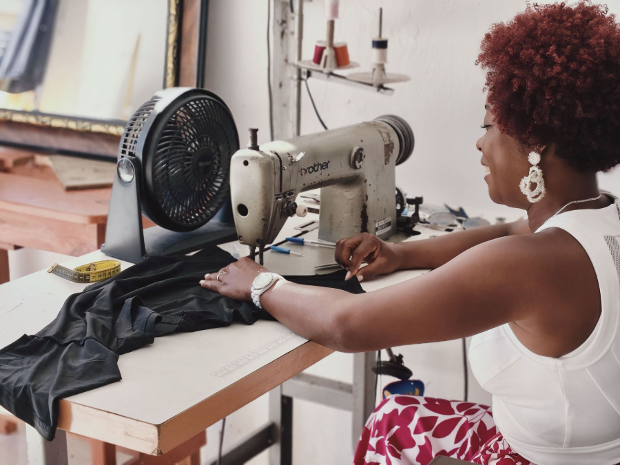 Person sewing black fabric at a machine in a workshop with a desk fan nearby