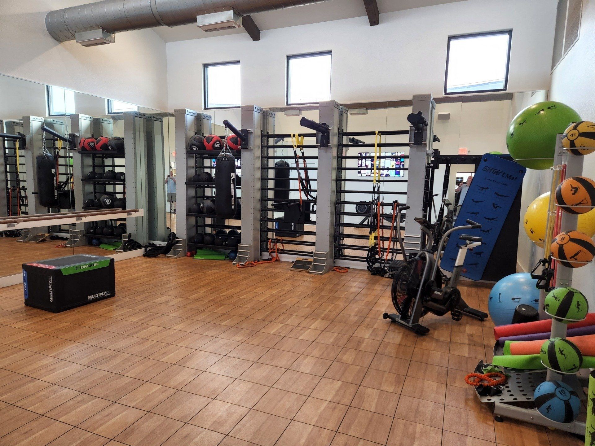 A bright, empty fitness studio with wooden flooring, exercise equipment racks, gym balls, and a stationary bike.