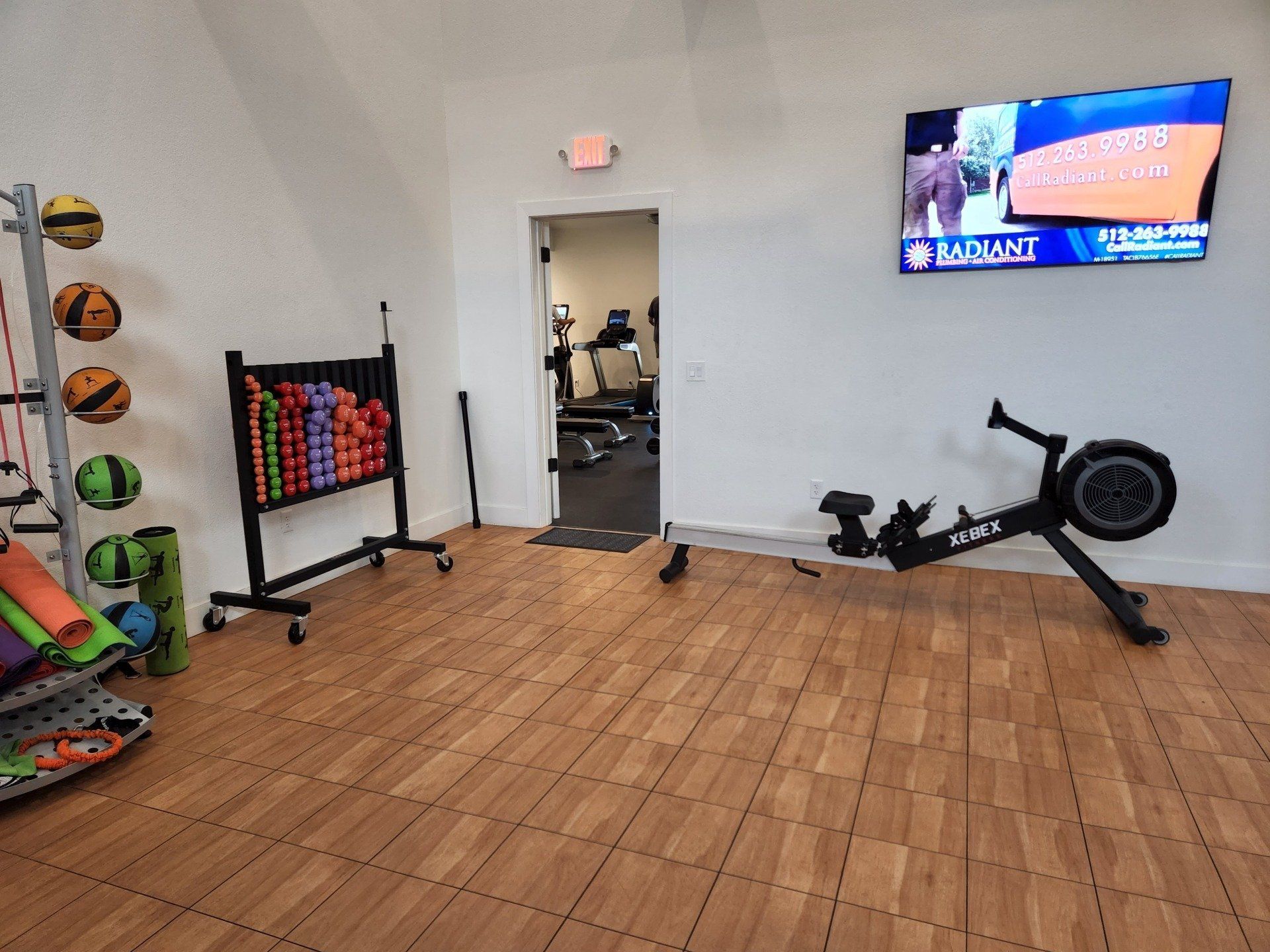 Small gym room with light wood floors, a dumbbell rack, a rowing machine, yoga mats, and a wall-mounted TV.