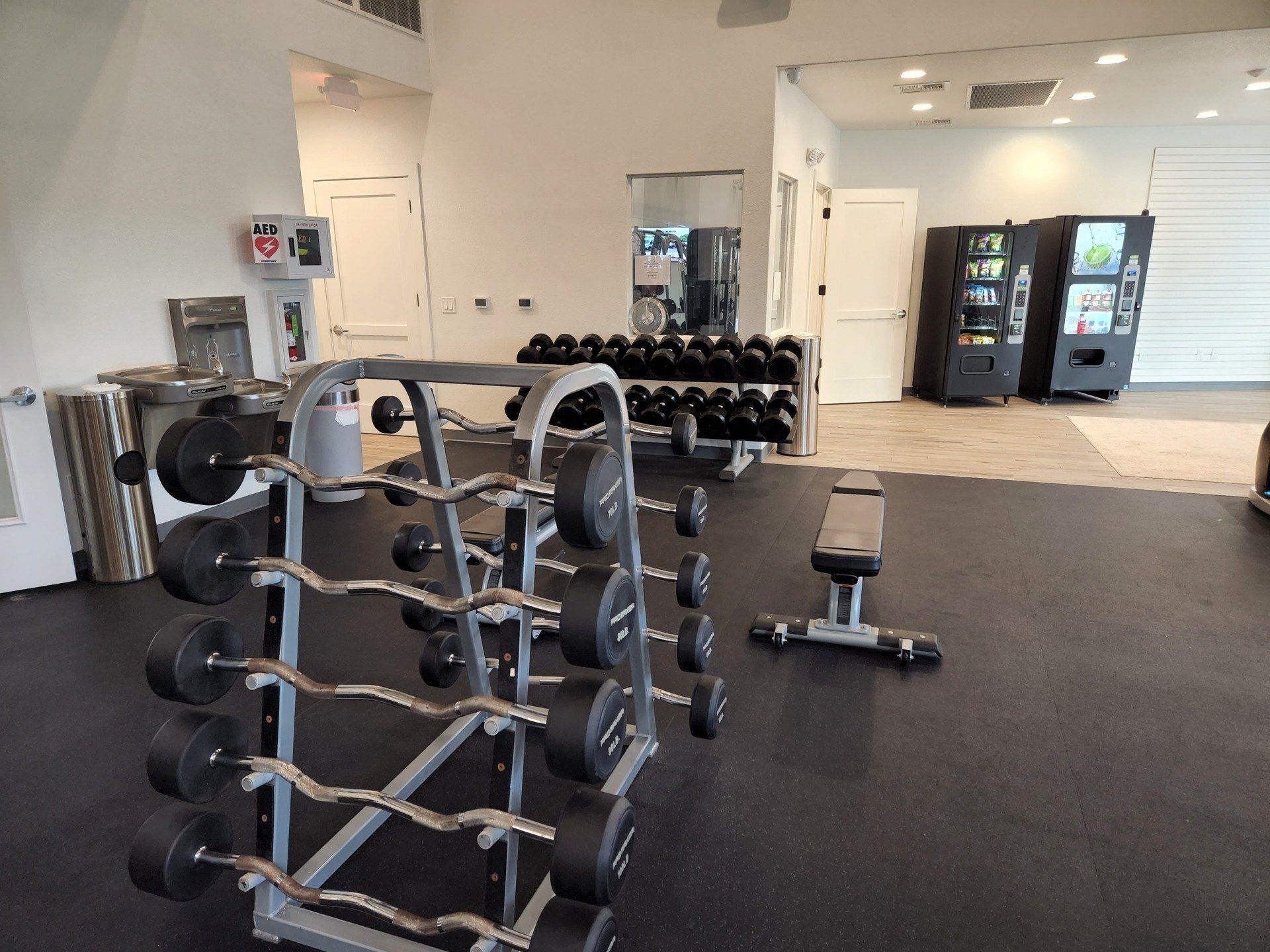 A gym with a barbell rack holding curved bars, a dumbbell rack in the back, a weight bench, and two vending machines.