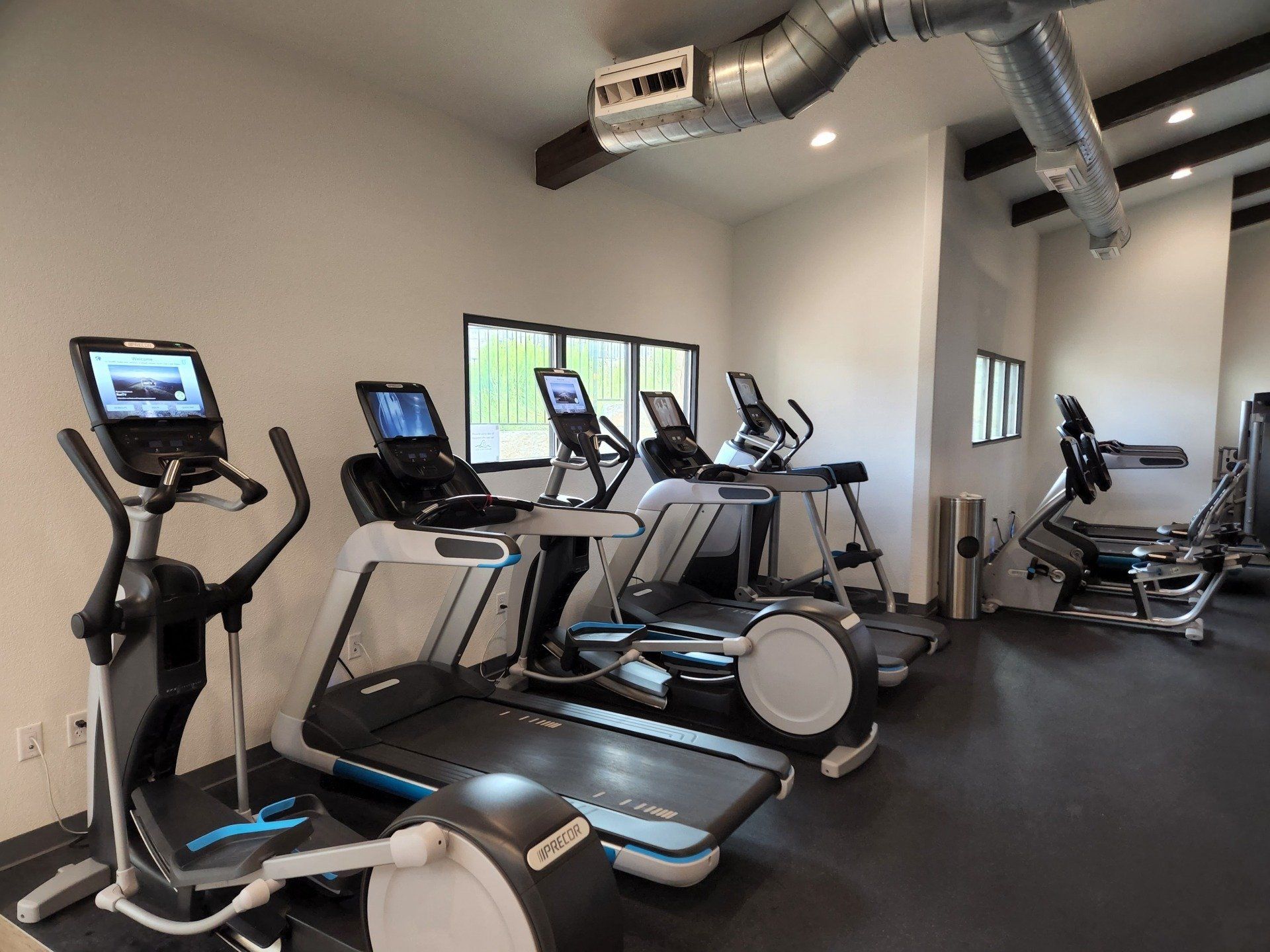 A brightly lit gym featuring a row of elliptical machines and treadmills with digital screens, situated on dark flooring.