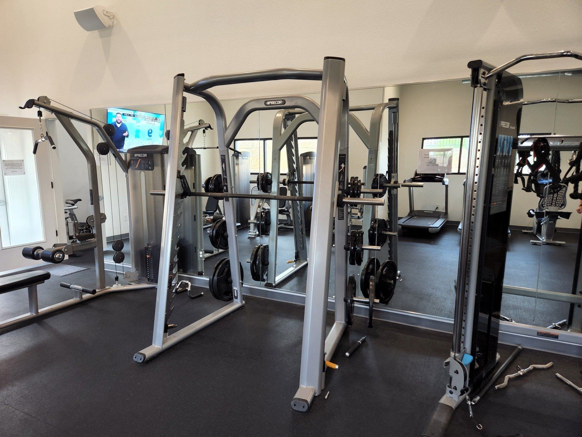 A clean, well-lit fitness center interior featuring a Smith machine in the center, free weights, and a treadmill.