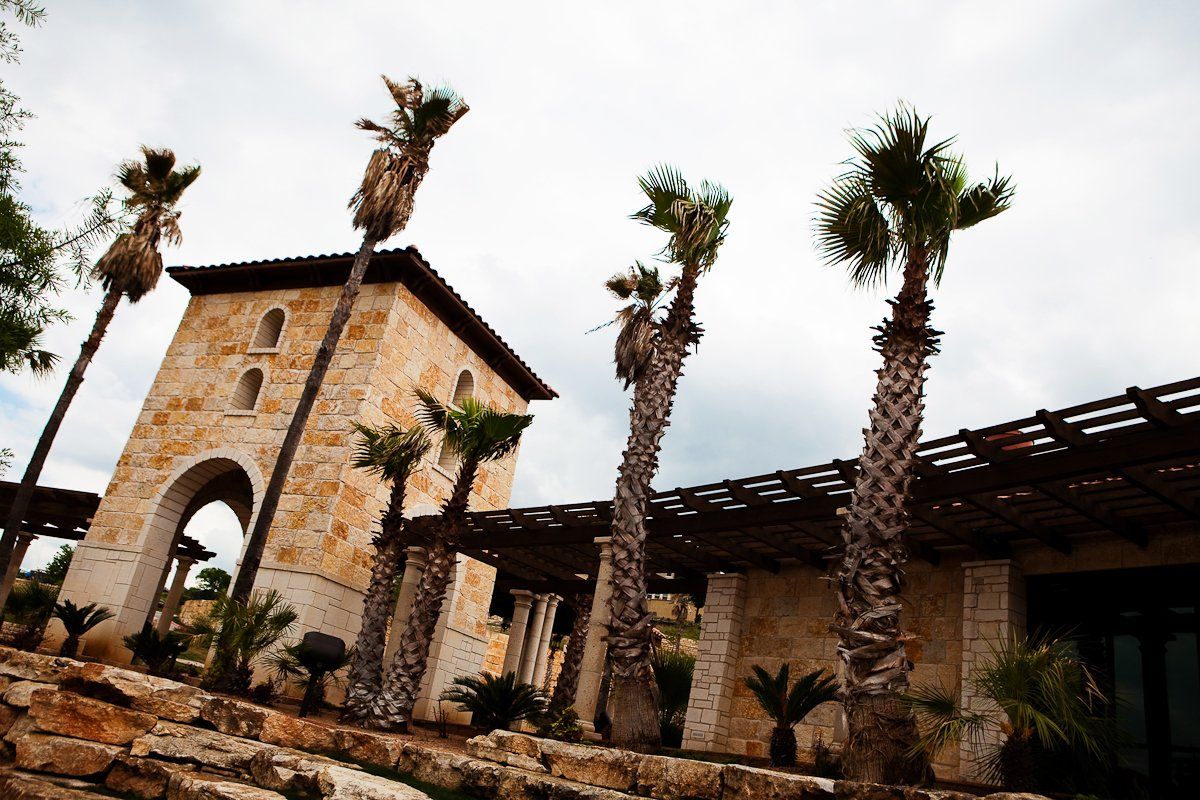 A stone building with a tall tower and a wooden pergola, surrounded by palm trees under a cloudy sky.