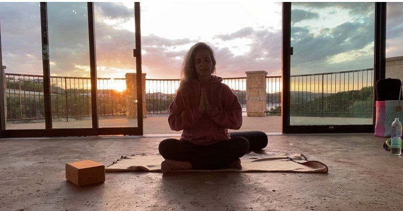A person sits in a meditation pose on a mat before large windows during a sunset.
