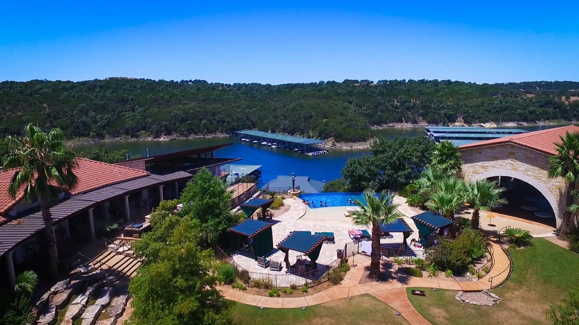 An elevated view of a resort pool area with cabanas, palm trees, and a lake with docks under a clear blue sky.
