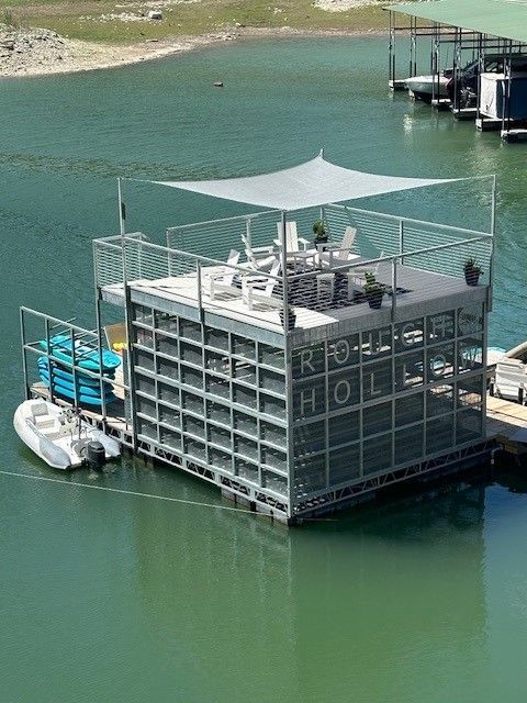 A gray, multi-level floating deck on a lake, featuring a top-level seating area under a shade sail and a small white boat.