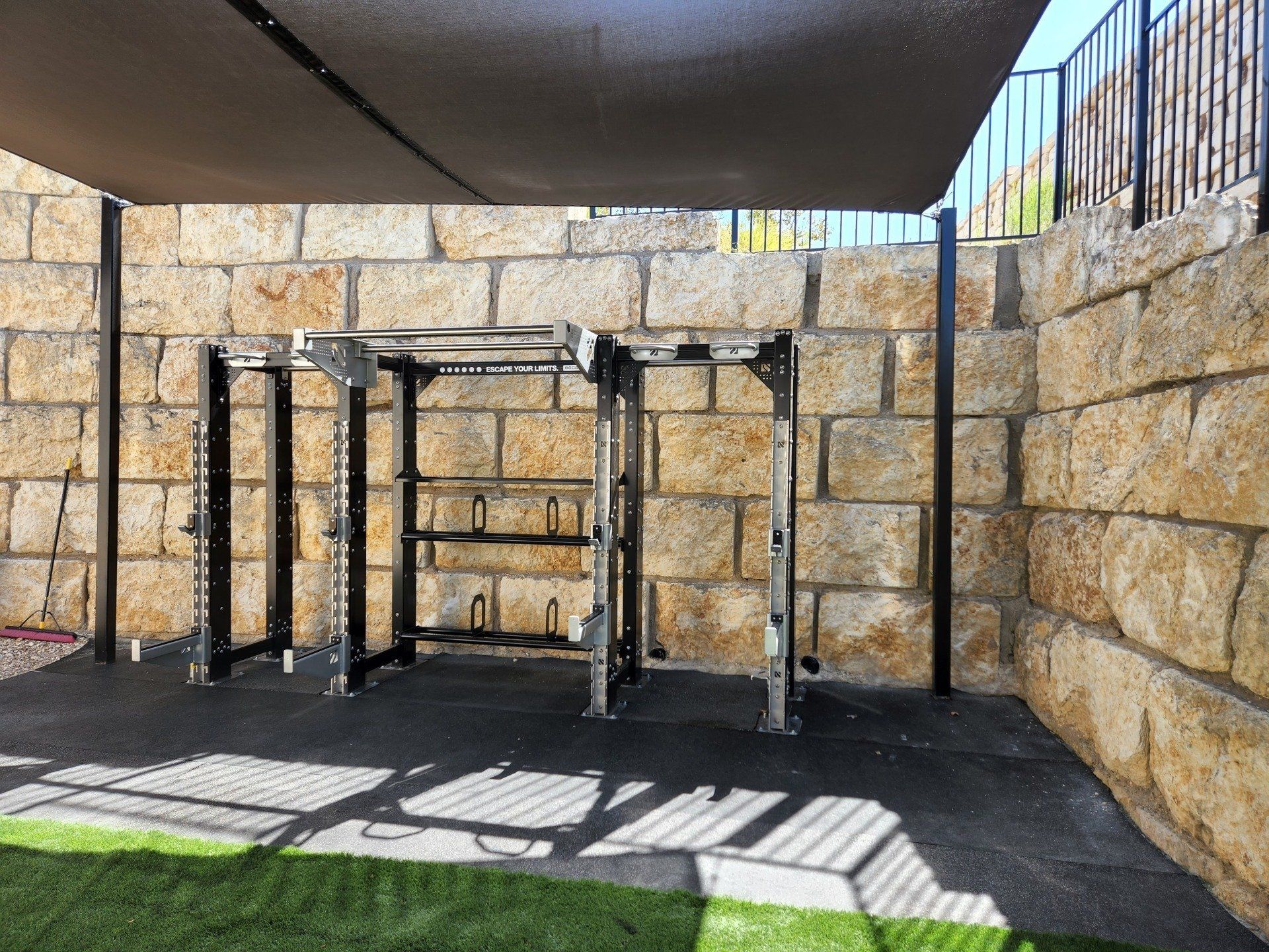 An outdoor gym area with a metal power rack structure on a rubber mat, backed by a natural stone retaining wall.