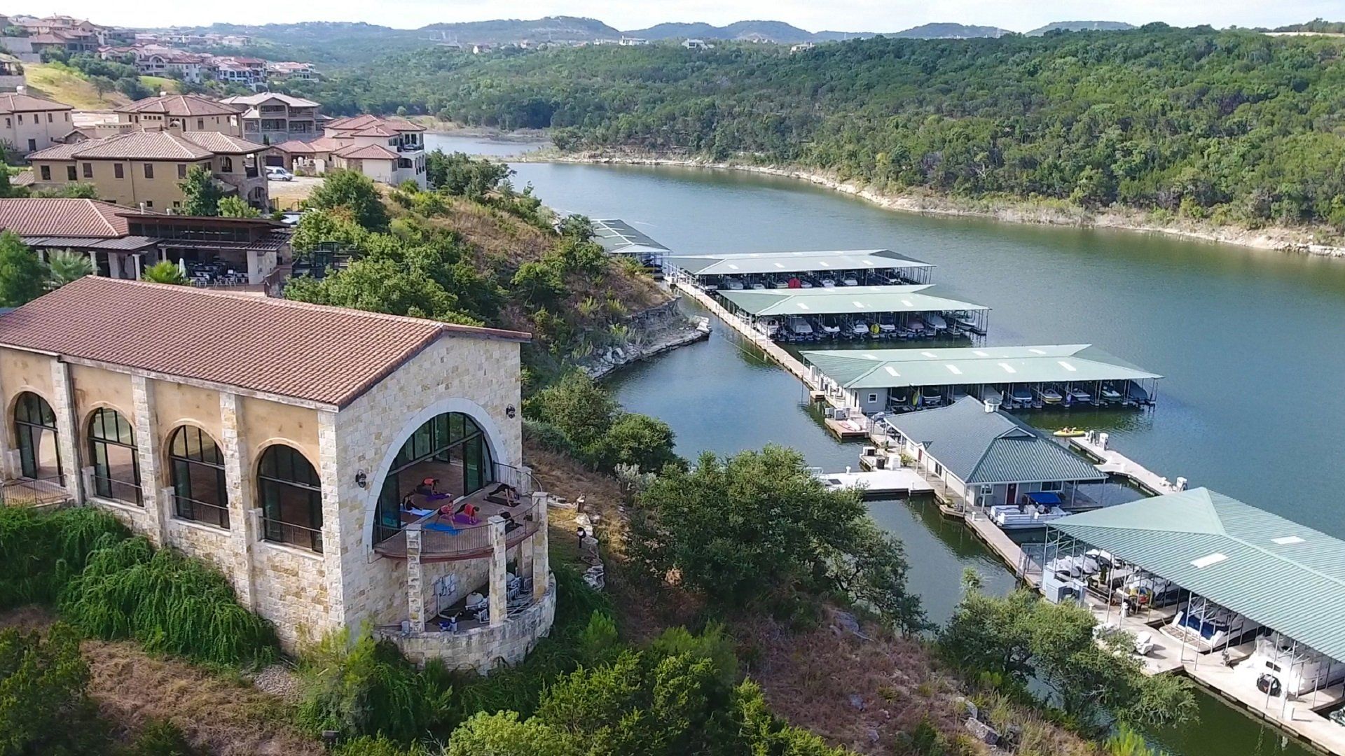 A stone building with arched windows overlooks a marina with several long, covered boat docks on a lake.