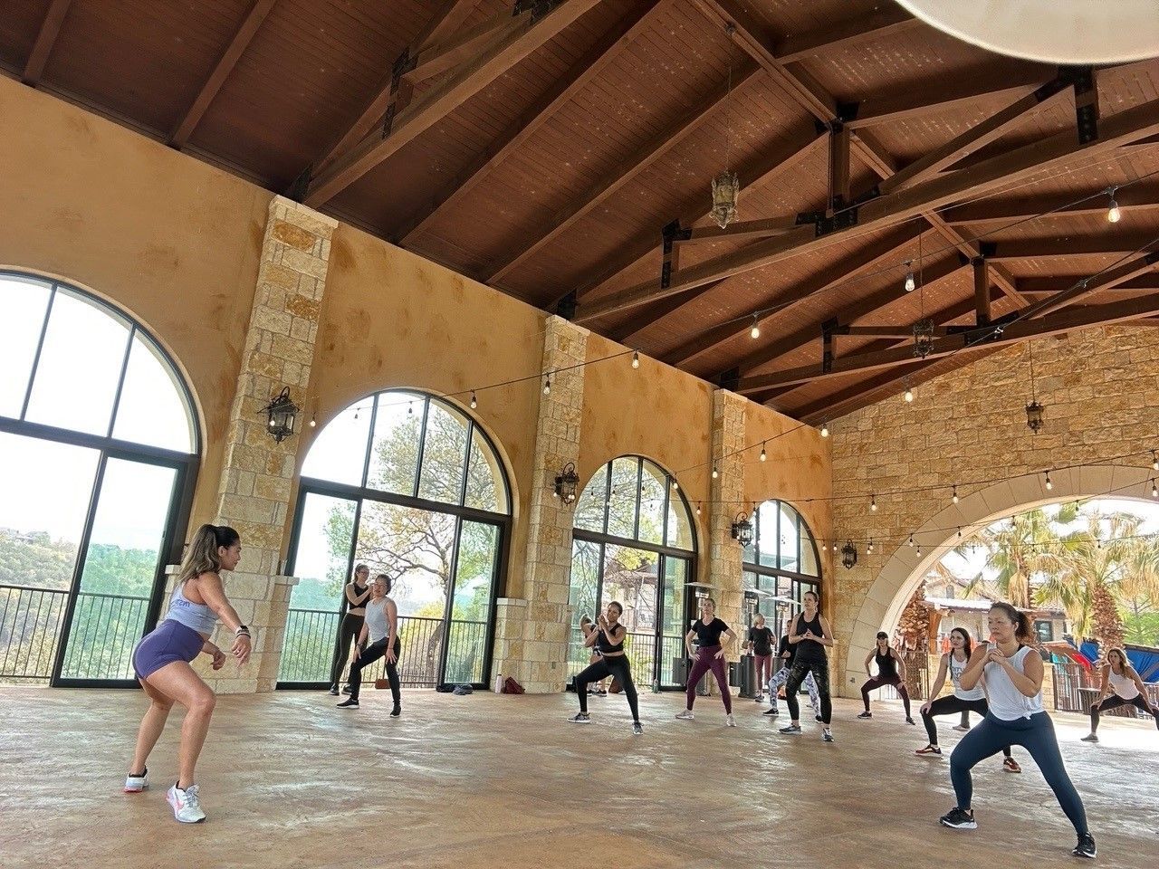 A group of people performs a fitness dance class in a spacious room with high arched windows and a wooden beam ceiling.