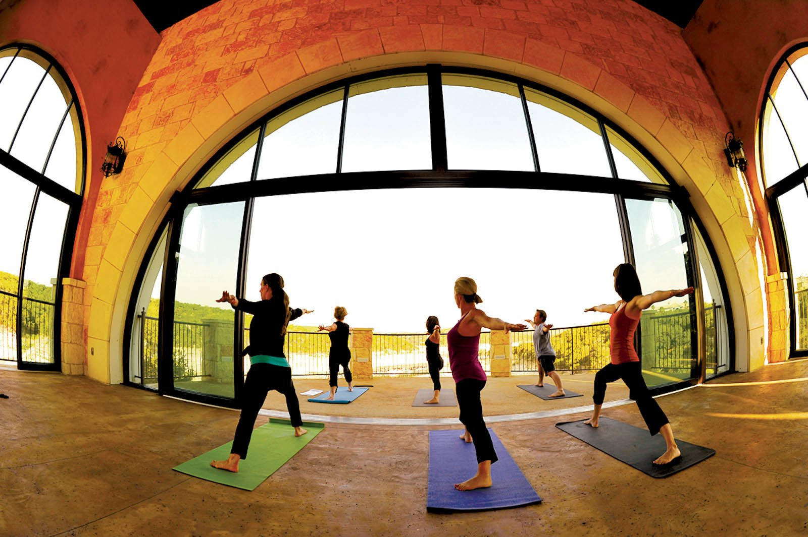Six people practicing yoga in a large, arched room with floor-to-ceiling windows overlooking a landscape.