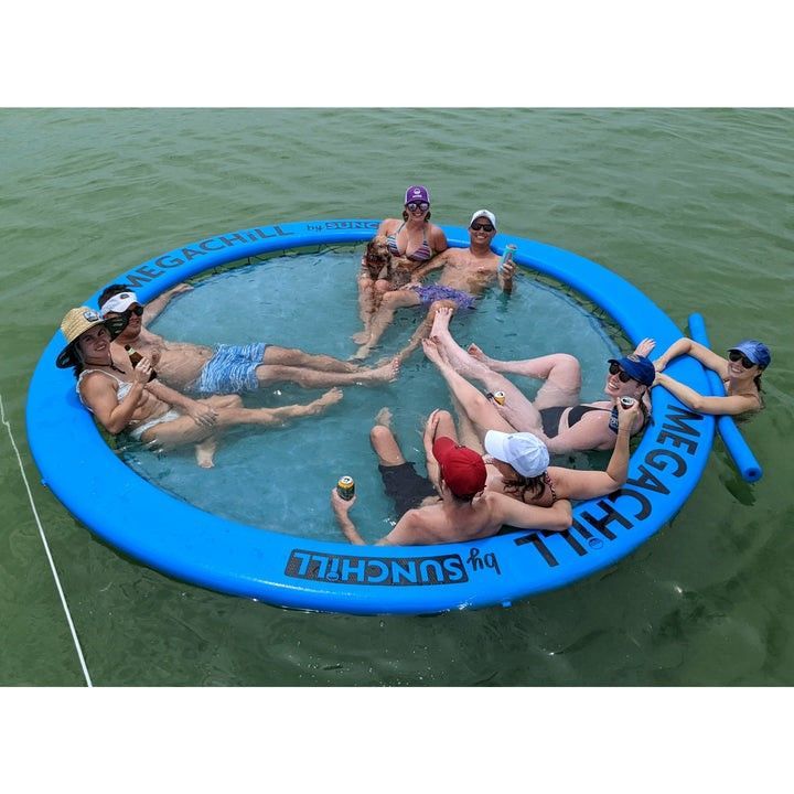 A group of people wearing hats and swimwear relax in a large, blue, circular inflatable pool floating on calm water.