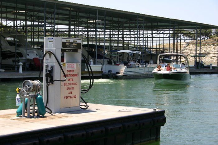 A fuel pump sits on a dock at a marina, with boats moored nearby under a covered structure and one boat approaching.