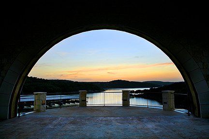 A stone archway overlooks a lake at sunset, with a metal railing and a serene, reflective water view.