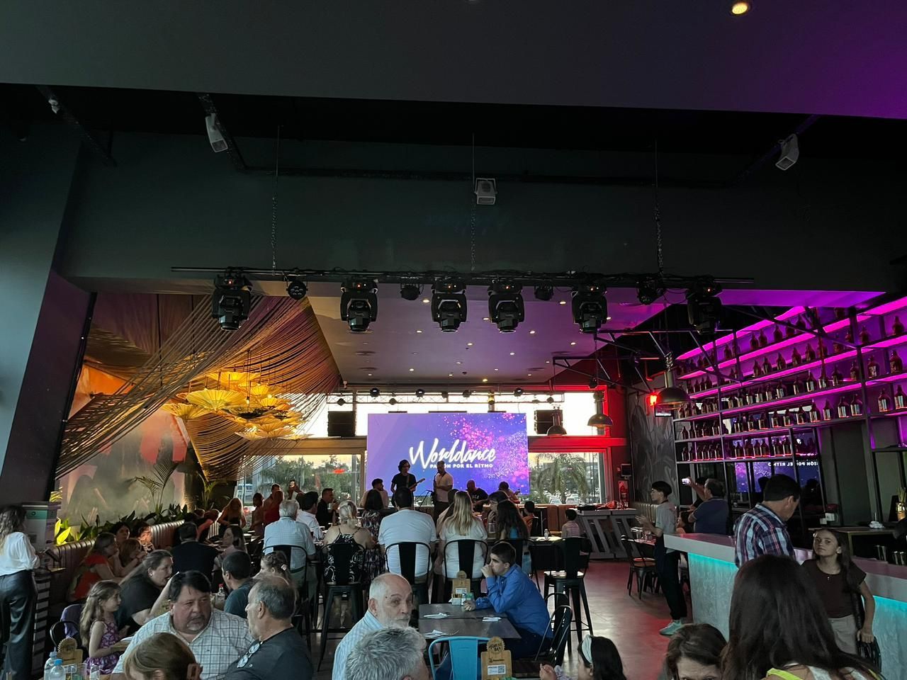 Interior de restaurante con multitud, escenario y bar, con iluminación violeta y pantallas grandes.