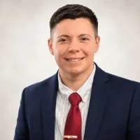 Portrait of a smiling person in a navy suit, white shirt, and red tie against a light background