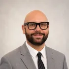 Headshot of a smiling man in glasses and a gray suit against a light background
