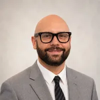 Headshot of a smiling man in glasses and a gray suit against a light background