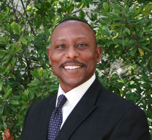 Smiling man in a dark suit and tie standing before leafy green trees
