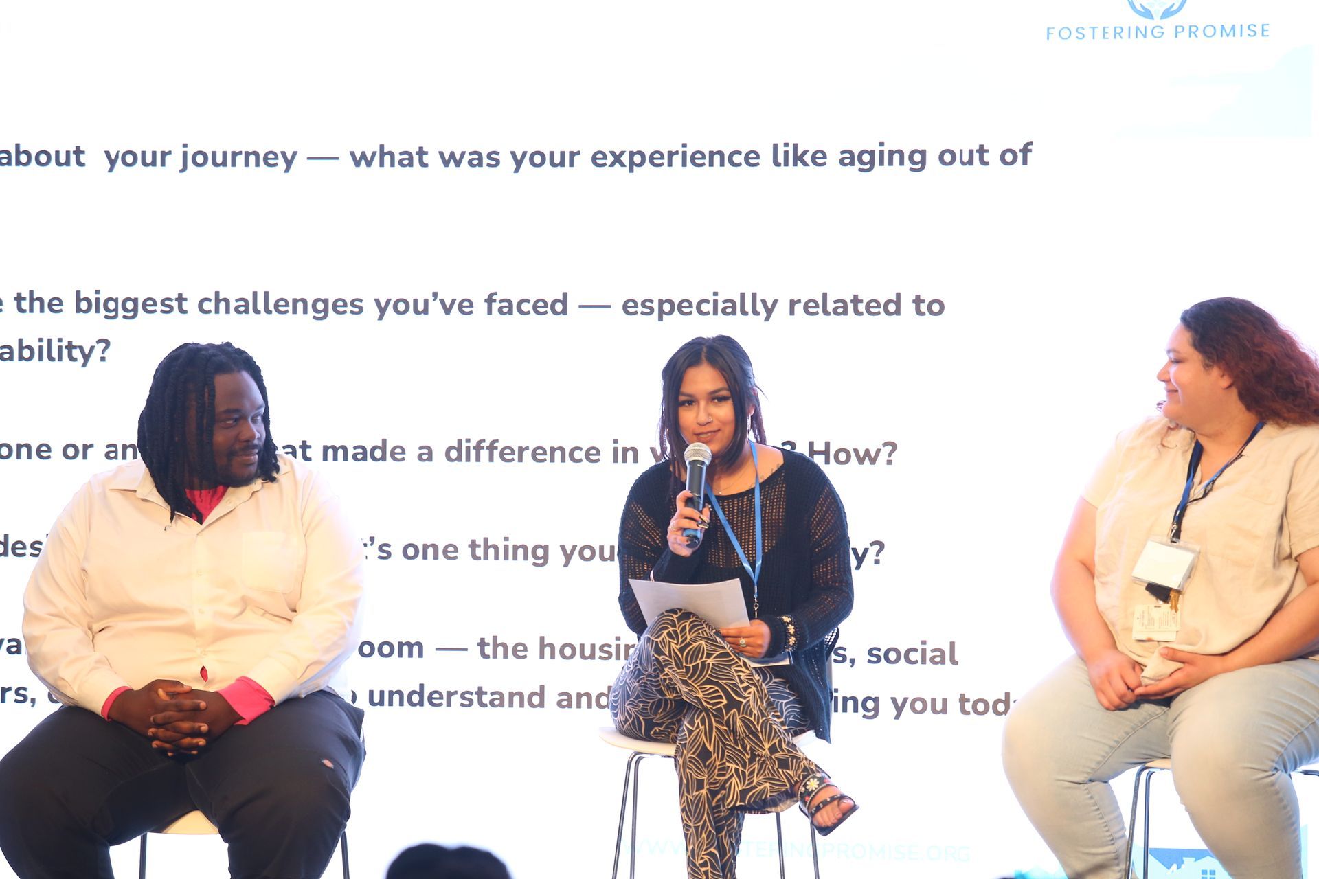 Three speakers seated on stage in a panel discussion before a text-covered backdrop.
