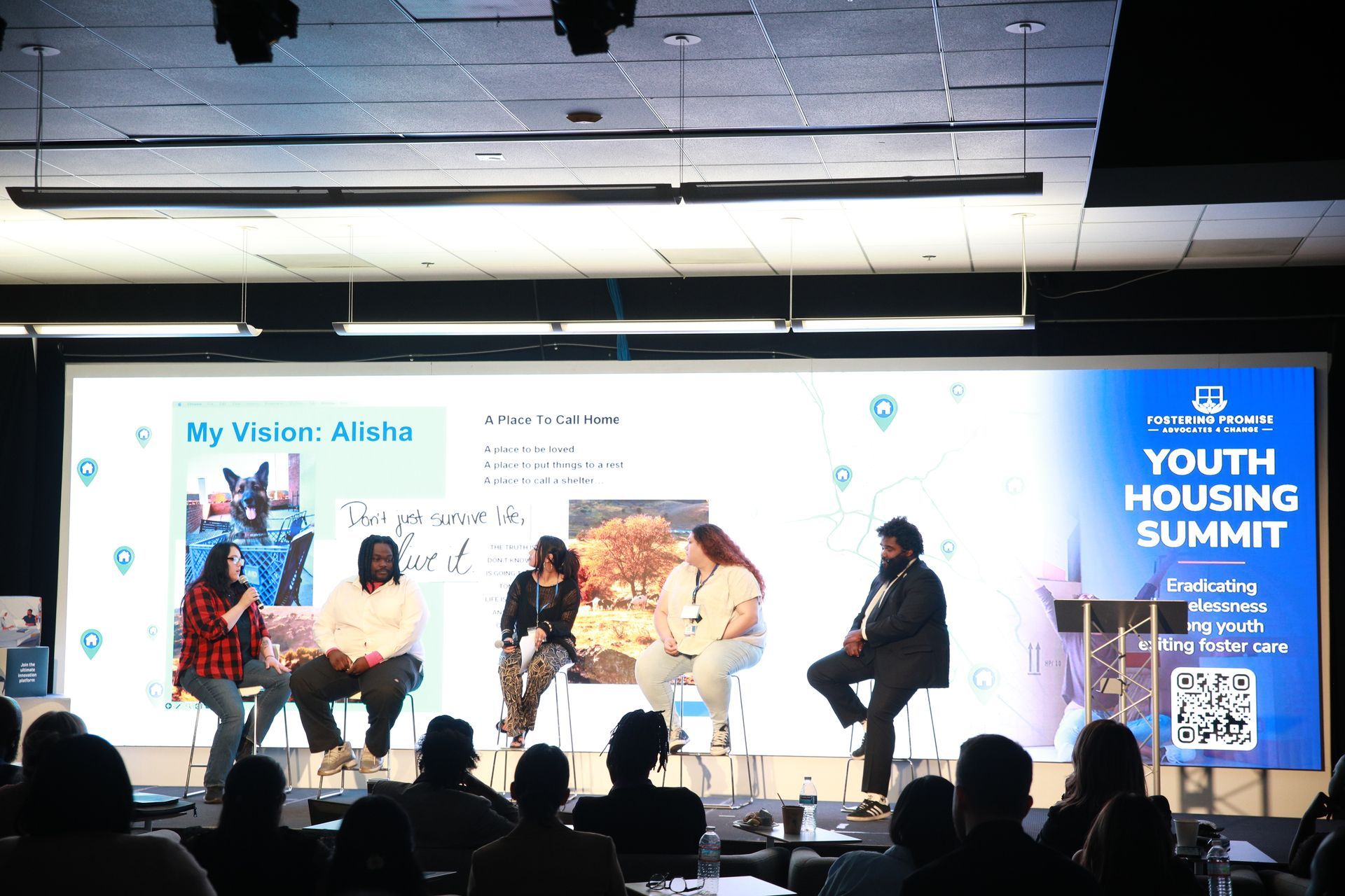 Panel discussion on stage at the Youth Housing Summit, with four seated speakers and blue event signage.