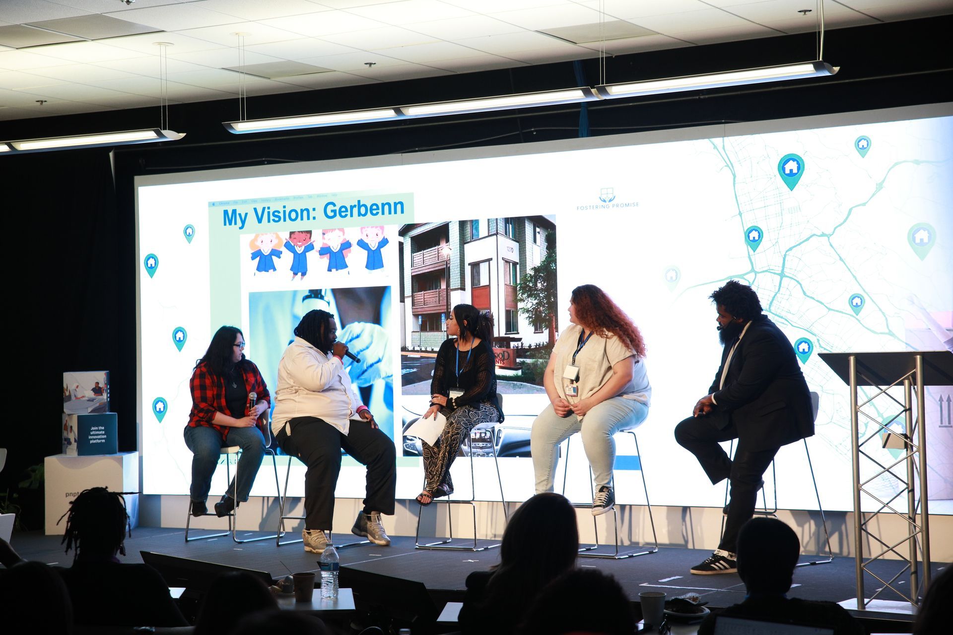 Panel discussion on stage with five seated speakers in front of a large screen backdrop.