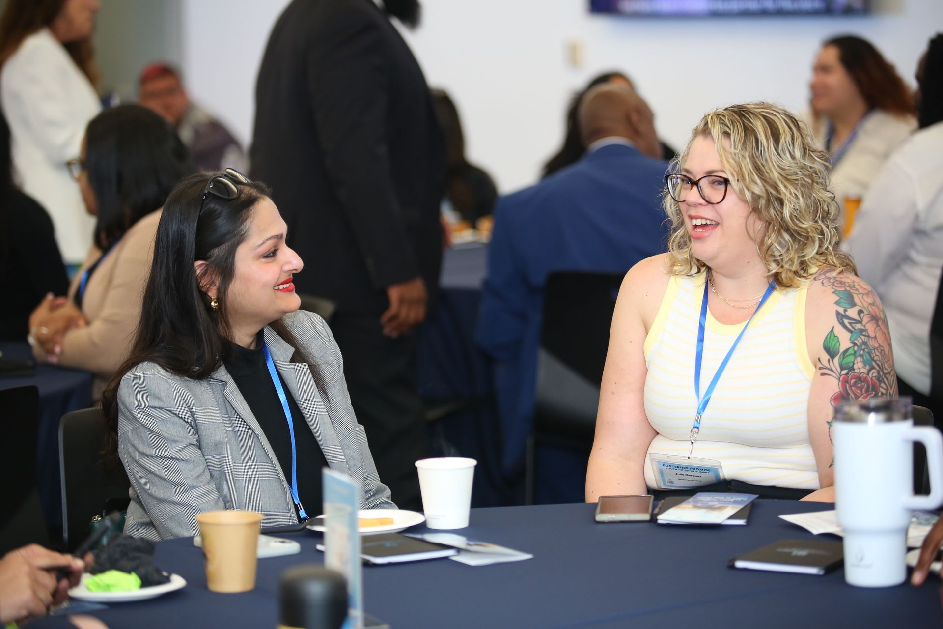 Two women chatting at a conference table with drinks and name badges in a bright event room